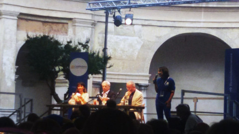 una donna e due uomini seduti a un tavolo su un palco davanti al pubblico